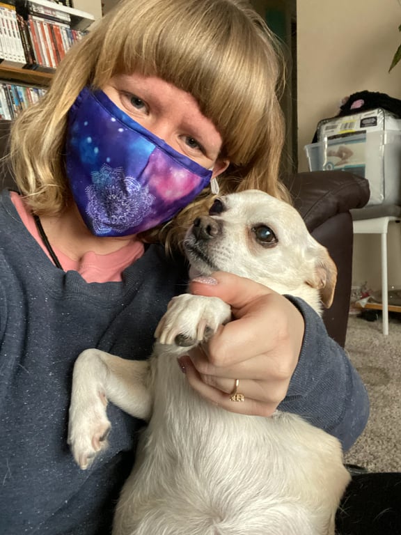 A woman with a galaxy face mask on holds the paw of a white chihuahua.